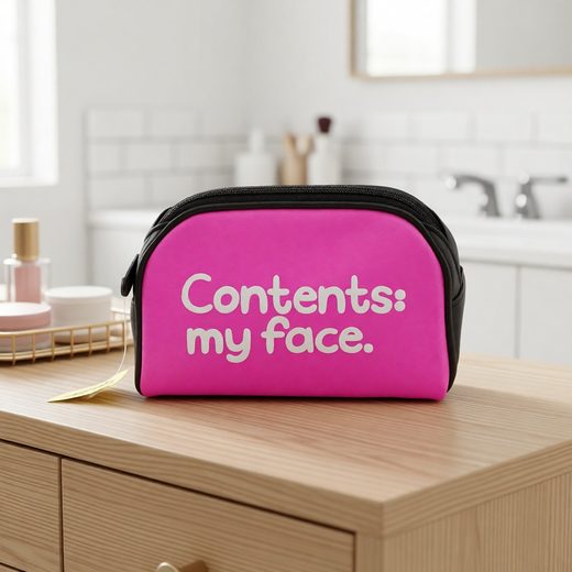 Vibrant pink zippered cosmetic bag with "Contents: my face" text on a light wood surface.