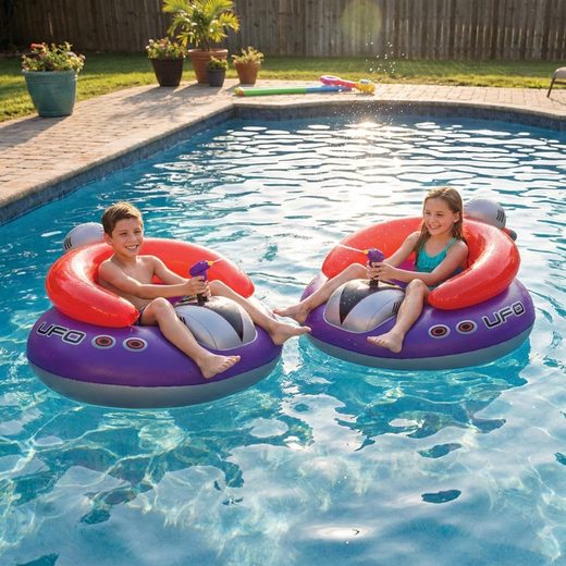 Two kids floating on purple UFO pool floats with water squirters in a backyard pool.